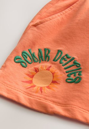 Tela de algodón naranja con texto bordado en verde "Deidades Solares" encima de un gráfico de sol con rayos amarillos y naranjas.