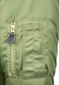 Žalios spalvos audinys su lygiu paviršiumi turi kišenę su užtrauktuku, kurio užtrauktukas auksinės spalvos, kišenę puošia siūlės šoninėje pusėje.