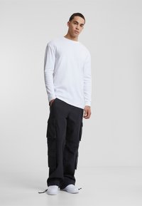 Hvit langermet skjorte, avslappet passform, kombinert med sorte cargo-bukser med flere lommer. Modellen har på seg hvite joggesko, plassert mot en ensfarget bakgrunn.