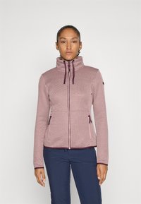 Kvinne iført en strukturert, mauve jakke med glidelås og snor, som st�år mot en simpel hvit bakgrunn, kombinert med marineblå bukser.