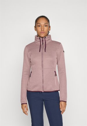 Frau in einer strukturierten, mauvefarbenen Jacke mit Reißverschluss und Kordelzügen, die vor einem schlichten weißen Hintergrund steht, kombiniert mit navyblauen Hosen.