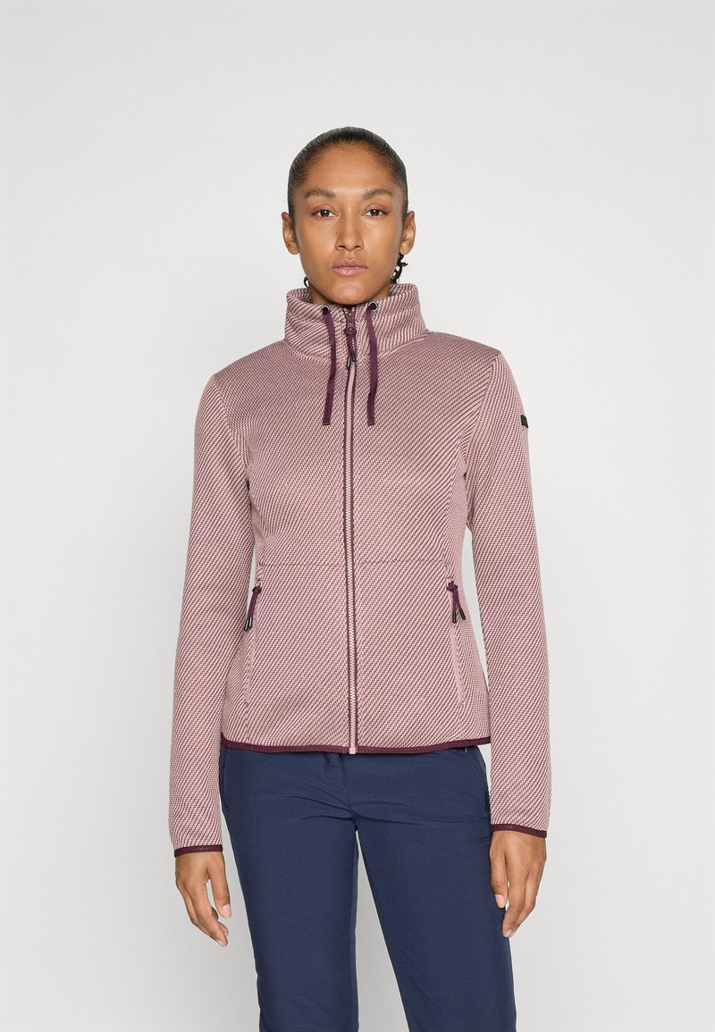 Kvinne iført en strukturert, mauve jakke med glidelås og snor, som står mot en simpel hvit bakgrunn, kombinert med marineblå bukser.
