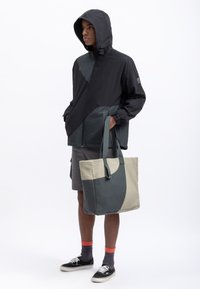 Lichte zwarte gewatteerde jas met groene accenten, gecombineerd met een grote, bicolor beige en donkergroene tote bag. Model draagt grijze shorts en sneakers.