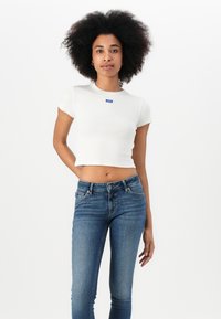 Fehér bordázott rövid ujjú crop top kék márkacímkével, karcsú kék farmernadrággal párosítva. Simább textúra és laza szabás.