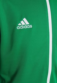 Giacca sportiva verde realizzata in tessuto liscio, con cerniera bianca, rifiniture bianche e un logo Adidas ricamato sul lato sinistro.