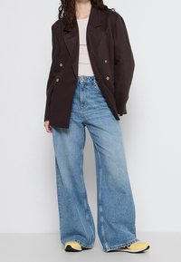 Femme portant un blazer marron foncé oversize, un haut blanc, un jean bleu clair à jambes larges et des baskets jaunes, debout devant un fond blanc uni.