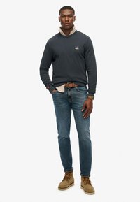 Man staat met zijn handen in zijn zakken, draagt een donker sweatshirt, een lichtbeige shirt, blauwe spijkerbroek, een bruine riem, een horloge en tan veterschoenen.