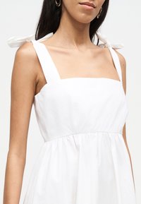 Γυναίκα που φοράει λευκό αμάνικο φόρεμα με δεμένους ιμάντες στους ώμους και χρυσά κρίκους στα σκουλαρίκια, εμφανίζεται από το στήθος μέχρι το πηγούνι.