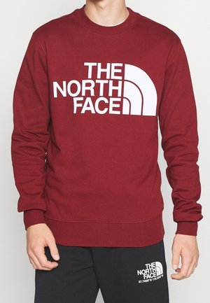 Person trägt einen kastanienbraunen Pullover und schwarze Hose, beide mit großen weißen "The North Face"-Logos auf der Vorderseite.