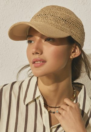 Young woman wearing a beige woven cap, striped shirt, and colorful beaded necklaces, looking slightly to the side against a light textured wall.