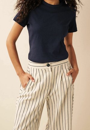 Kobieta w granatowym, krótkim rękawem t-shircie włożonym w białe spodnie z wysokim stanem z czarnymi pionowymi paskami, z rękami w kieszeniach.
