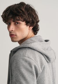 Grijze rits hoodie met een zachte textuur en een capuchon, getoond op een model dat iets wegdraait, zodat het ontwerp van het kledingstuk goed zichtbaar is.