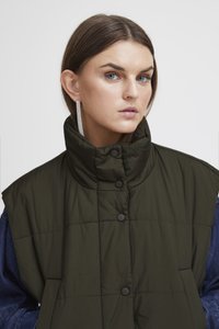 Tumedast rohelisest padded vestist kõrge krae ja mustade neetidega. Omab tikitud tekstuuri ja laia lõiget, mille peal on denimist särk.