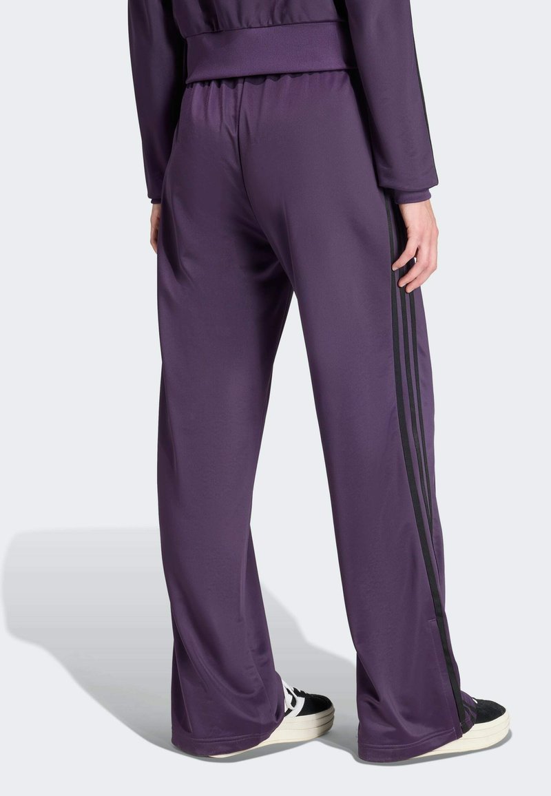 Pantalon de sport violet avec une texture brillante, featuring des rayures noires sur les côtés, une taille élastique et une coupe décontractée, évasée.