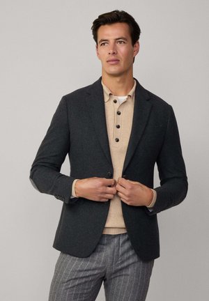 Blazer gris foncé avec revers, fermeture à un bouton, porté sur un polo beige à côtes. Pantalon à rayures grises visible en dessous.