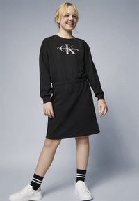 Vestido de sweatshirt preto com cintura elástica. Apresenta o logo "Calvin Klein Jeans". Combinado com ténis brancos e meias pretas listas.