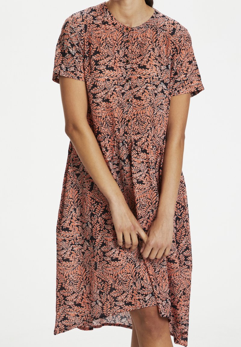 Femme portant une robe à manches courtes avec boutons sur le devant, motif de feuilles roses et noires, debout, les mains doucement jointes devant.