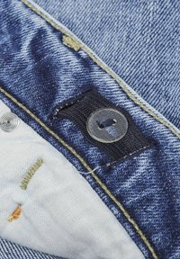 Denimtyg i ljus och mörkblå nyanser med kontrasterande gul sömnad, med en metallknapp och texturerade tygdetaljer.