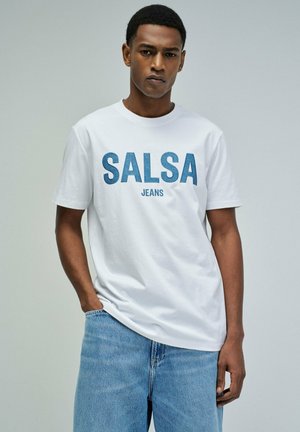 Hombre con camiseta blanca con texto "SALSA JEANS" y vaqueros azul claro, de pie con una mano en el bolsillo contra un fondo liso.