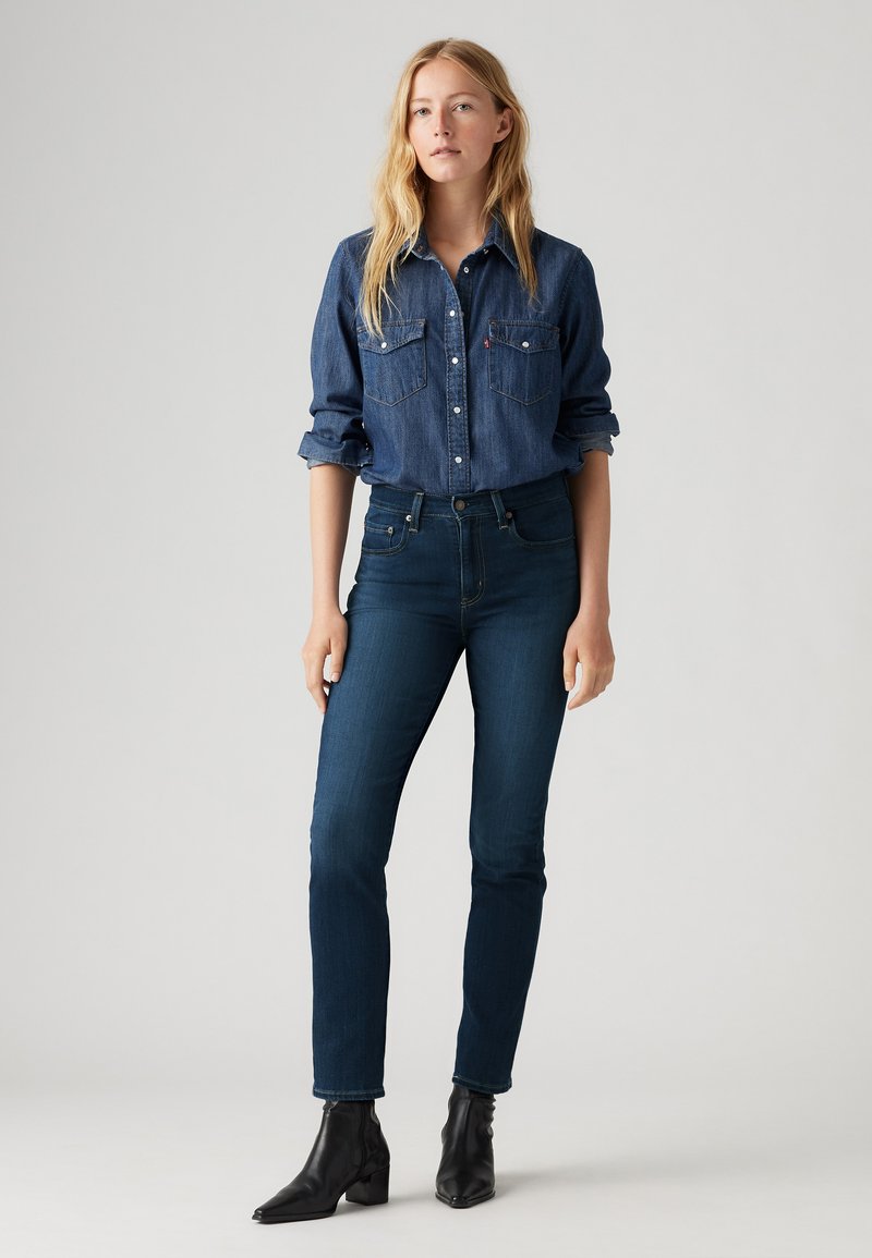 Chemise en denim avec fermeture à boutons, deux poches poitrine et manches retroussées ; portée avec un jean skinny bleu foncé et des bottines noires.