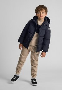 Jūras spēku pūkains mētelis ar kapuci, bēšs sporta krekls un bēšas sporta bikses. Melni sporta apavi ar baltām akcentiem. Gludas tekstūras, ikdienišķs dizains.