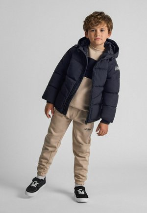 Jūras spēku pūkains mētelis ar kapuci, bēšs sporta krekls un bēšas sporta bikses. Melni sporta apavi ar baltām akcentiem. Gludas tekstūras, ikdienišķs dizains.