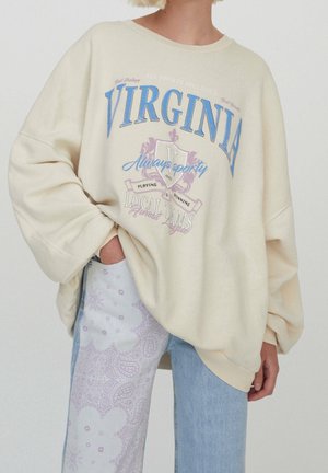 Osoba nosząca oversize kremową bluzę z napisem ""VIRGINIA"" oraz niebieskie dżinsy z jasnofioletowym wzorem paisley na jednej nogawce.