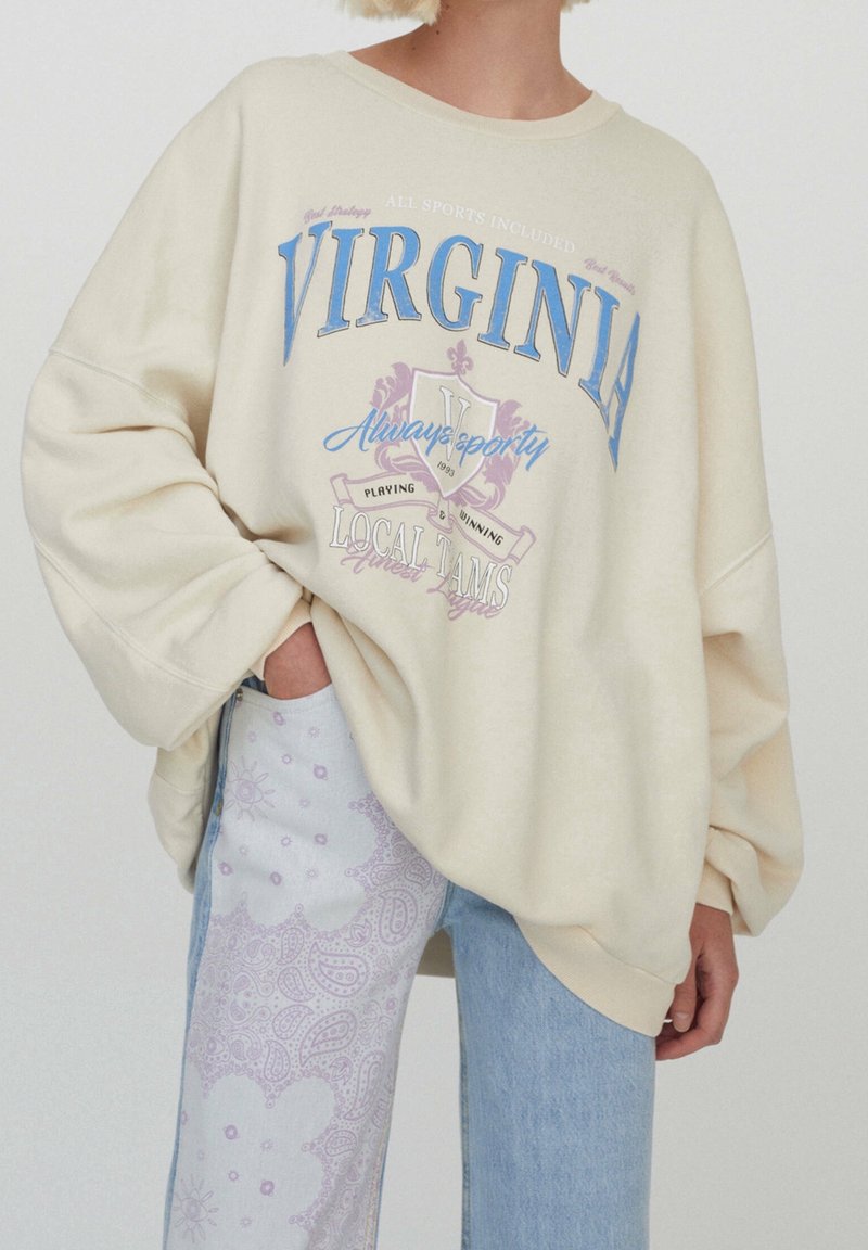 Personne portant un sweat-shirt crème oversize avec le texte ""VIRGINIA"" et un jean bleu avec un motif cachemire violet clair sur une jambe.