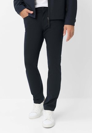 Mand iført sorte slim-fit bukser, hvide sneakers og en mørk jakke, står med den ene hånd i lommen mod en hvid baggrund.
