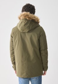 Hombre vestindo una parka verde oliva con capucha forrada de piel, de pie de espaldas, combinada con jeans azules sobre un fondo blanco.