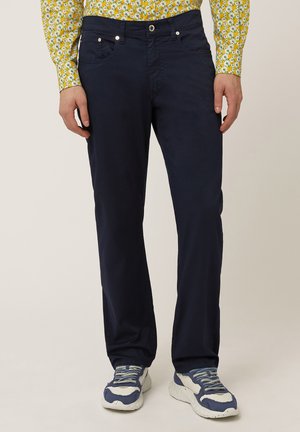 Pantalones de algodón azul marino con corte recto, bolsillos frontales y herrajes plateados. Combinados con una camisa amarilla con estampado y zapatillas deportivas.