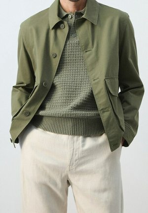 Uomo che indossa una giacca verde oliva con bottoni sopra un maglione verde lavorato a maglia con pantaloni color crema, mani in tasca.