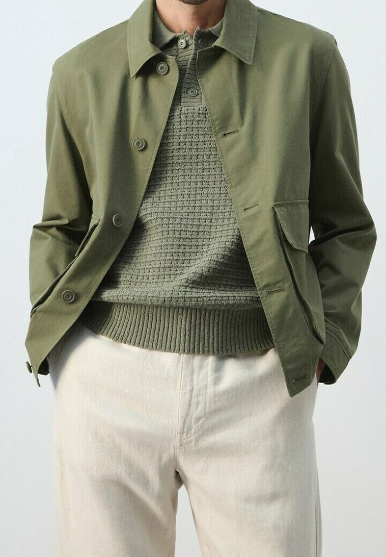 Homme portant une veste boutonnée vert olive par-dessus un pull en maille verte texturée avec un pantalon couleur crème, les mains dans les poches.