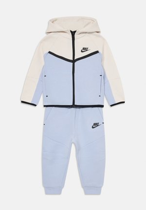 Dušo dalies Nike vaikų treniruočių kostiumas su šviesiai mėlynomis kelnėmis ir grietinėlės spalvos džemperiu su juodais apvadais ir logotipais, išsidėstęs ant balto fono.