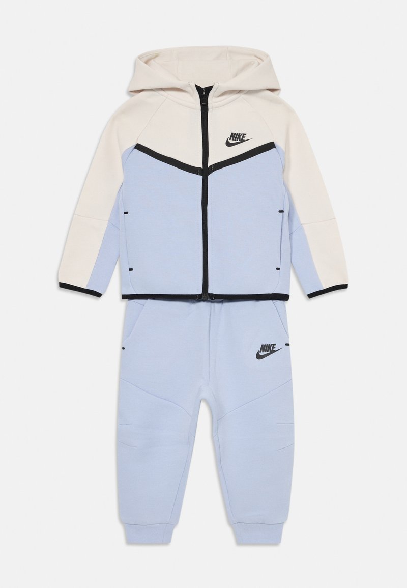 Dušo dalies Nike vaikų treniruočių kostiumas su šviesiai mėlynomis kelnėmis ir grietinėlės spalvos džemperiu su juodais apvadais ir logotipais, išsidėstęs ant balto fono.