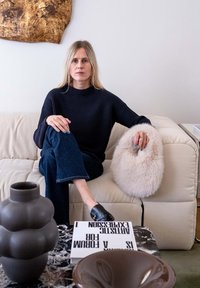 Maglione lavorato a maglia blu scuro, jeans blu, scarpe scure, con un cuscino morbido di colore crema in mano. Vaso scuro e libri su un tavolo di marmo.