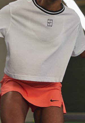 Personne portant un t-shirt Nike court blanc avec des rayures noires au col et un short de sport Nike orange vif, en pleine action sur un terrain de sport.