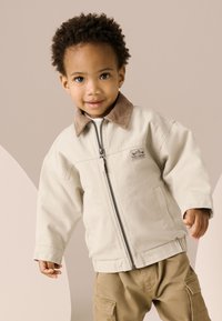 Bézs cipzáras dzseki barna kordbársony gallérral, hosszú ujjakkal és oldalsó zsebbel, khaki színű cargo nadrág fölött viselve.