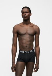 Αθλητικός μαύρος άνδρας στέκεται μπροστά σε λευκό φόντο φορώντας μαύρα boxer Tommy Hilfiger με λογότυπο στη μέση.