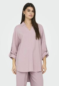 Blouse rose clair, ample, avec un col en V, des manches retroussées à trois quart et un ourlet légèrement incurvé. Tissu doux et texturé.