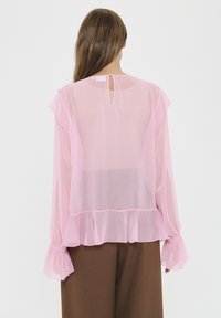 Rosa transparente Bluse mit Rüschensleeves, Rückenschlitz und fließendem Saum. Kombiniert mit braunen Hosen, die eine mehrschichtige Textur zeigen.