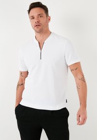 Bílý polo triko s krátkým rukávem a límcem na čtvrtinu zipu, texturovaná látka. Nosit s černými kalhotami. Obsahuje tetování na levém rameni.