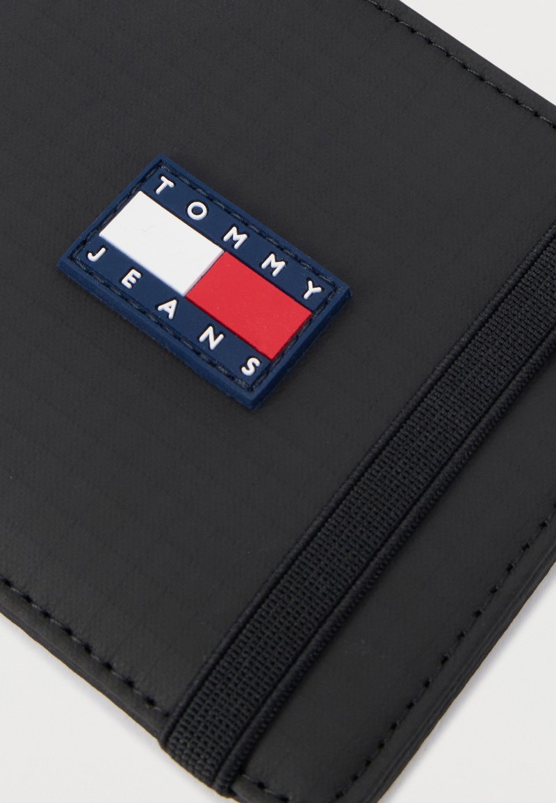 Portafoglio nero texturizzato con una toppa rettangolare cucita in blu, bianco e rosso con la scritta "TOMMY JEANS" in rilievo.