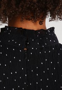 Black, textured fabric with white polka dots. Features a ruffled neckline, button closure at the back, and subtle pleating details.