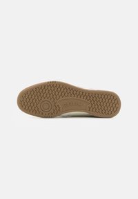 Reebok sneaker talpa érdes barna gumis felülettel. Kör alakú logó és mintázott tapadás a jobb érintkezésért. A fehér felsőrész az élnél látható.
