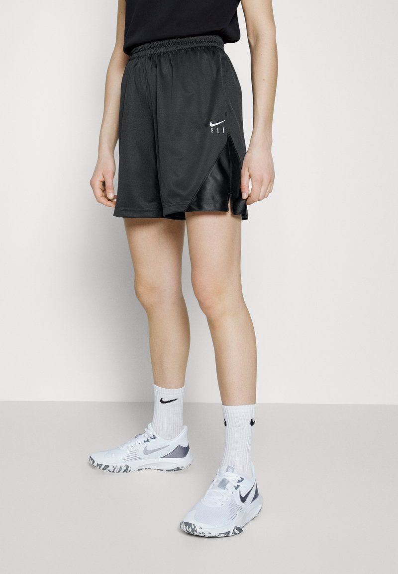 Zwarte sportshorts van mesh stof met zijsplitten, voorzien van een wit Nike-logo. Gecombineerd met witte sneakers en kousen tot aan de enkel.