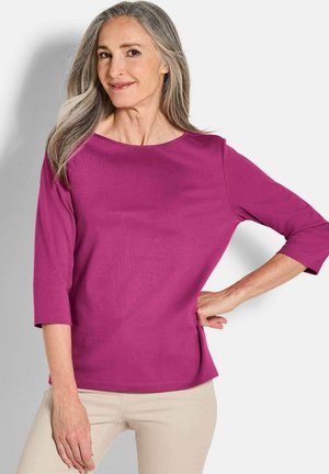 Magenta Langarmshirt mit weitem Ausschnitt und seitlichen Schlitzen, aus glattem Stoff gefertigt. Kombiniert mit hellbeigen Hosen.
