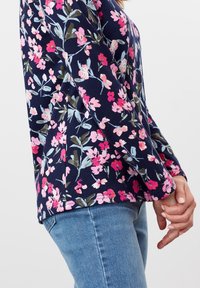 Námořnická květinová košile s dlouhým rukávem, která se vyznačuje růžovými, bílými a zelenými květy na jemném materiálu; kombinovaná se světle modrými džíny.