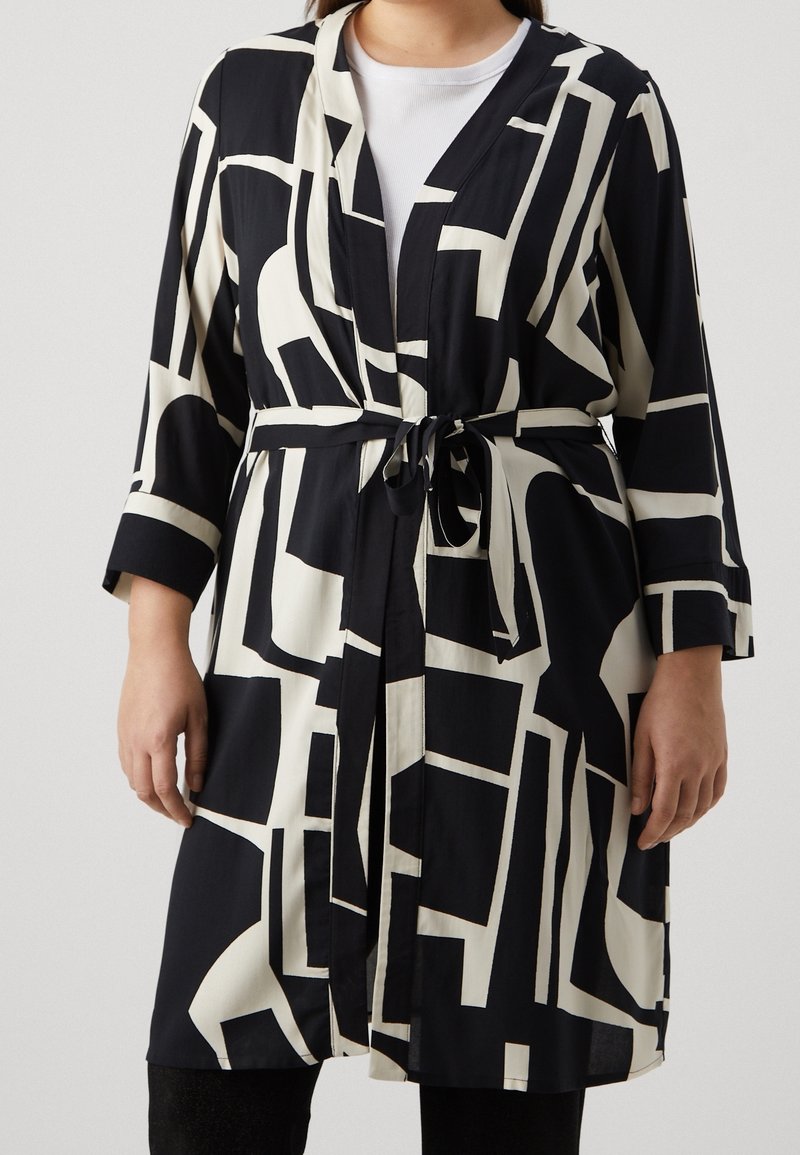 Femme portant une robe à motifs géométriques noir et blanc avec une ceinture à la taille, par-dessus une chemise blanche et un pantalon noir.