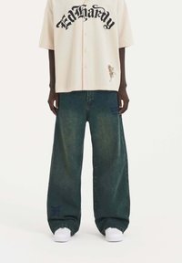 Košile s dlouhým rukávem v krémové barvě s černým logem "Ed Hardy" a květinovou výšivkou; volné tmavě zelené džíny; bílé tenisky s texturovanou podrážkou.